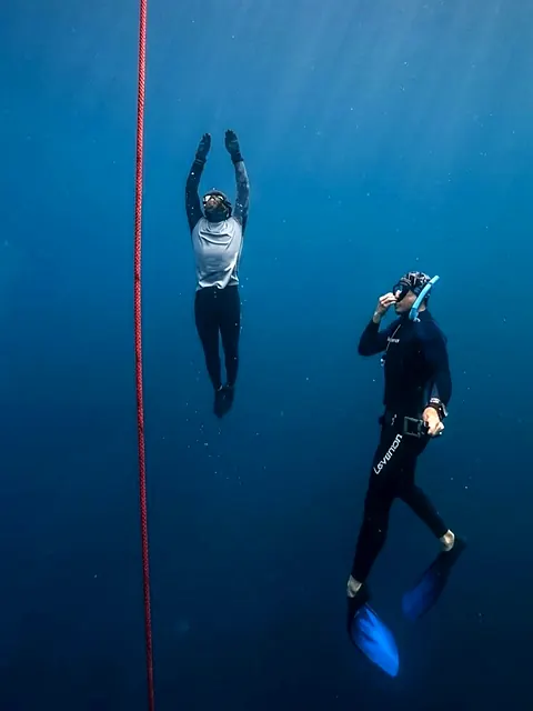 Dos apneístas suspendidos en el azul junto a una cuerda roja, uno con los brazos arriba