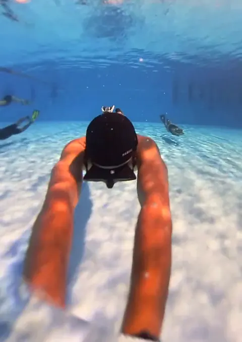 Un apneísta de gorro negro se desliza con los brazos extendidos hacia el fondo claro de la piscina, con otros makaras nadando más allá.
