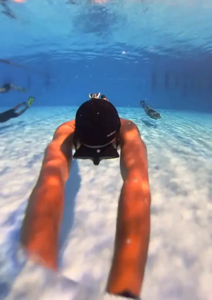 Un apneísta con gorro negro se desliza hacia el fondo de la piscina; arriba, la superficie refleja un cielo rosado